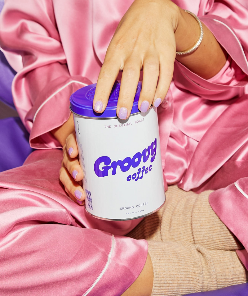 Person holding a can of 'Groovy Coffee' in pink satin pajamas.