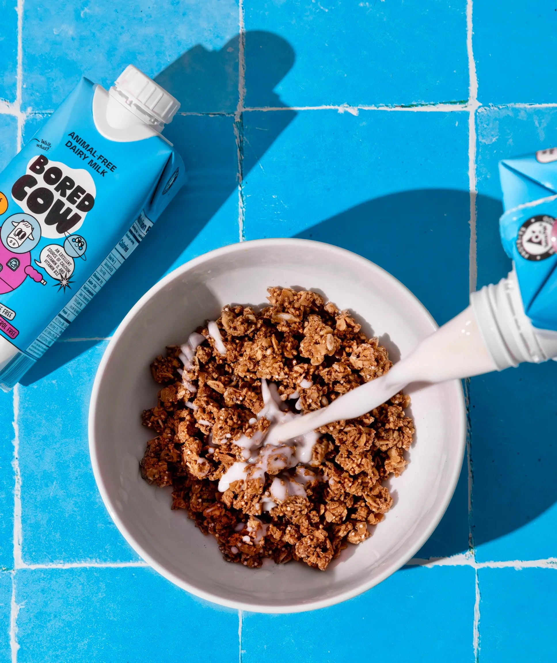 Bowl of cereal with milk being poured, Bored Cow milk cartons on a blue tiled background