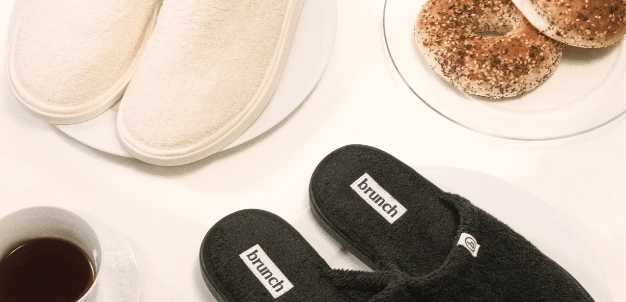 Pair of black slippers with 'brunch' branding on a white surface with coffee and bagels.