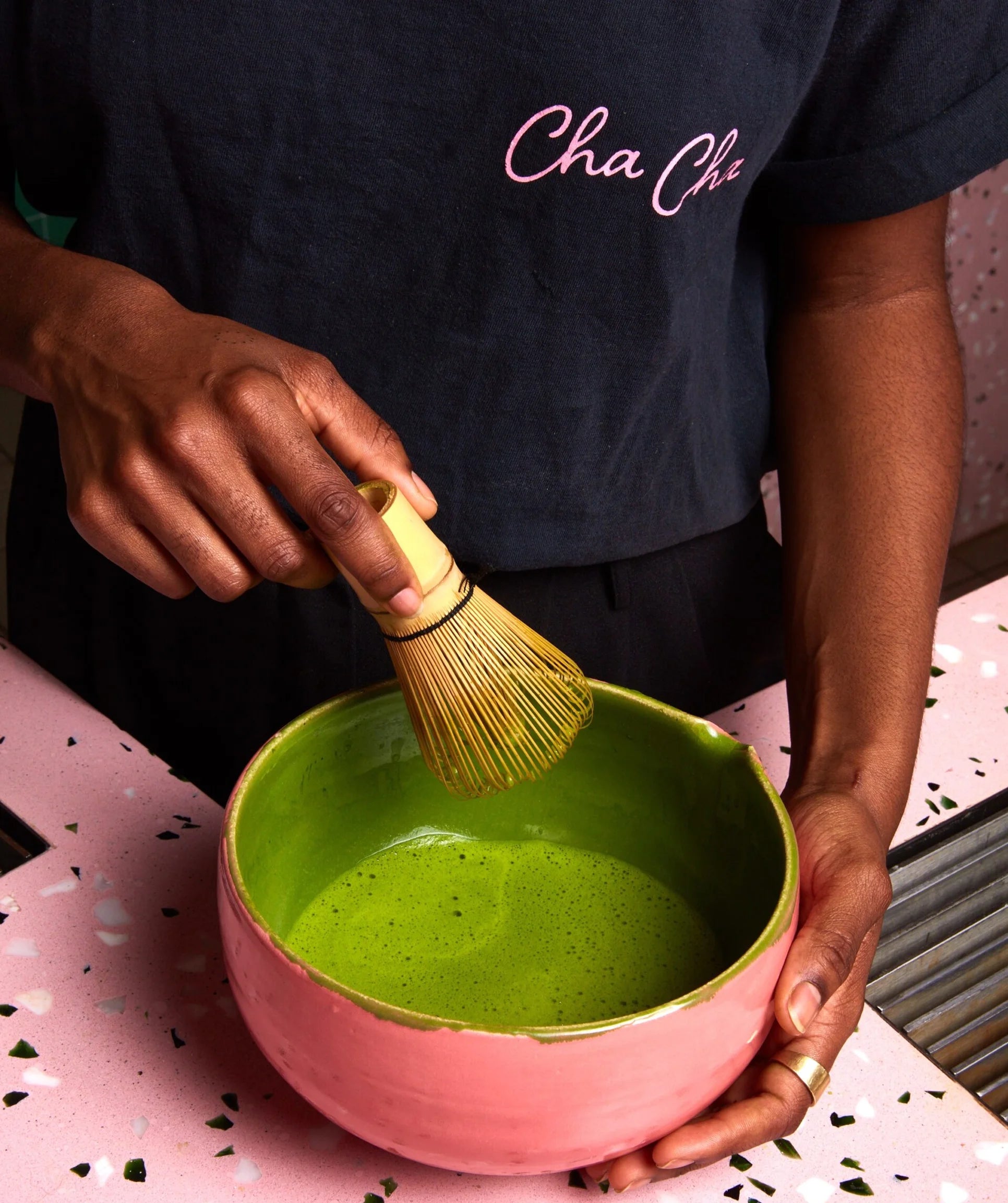 Person stirring green tea in a pink bowl with a whisk, wearing a black shirt with 'Cha Cha' text.
