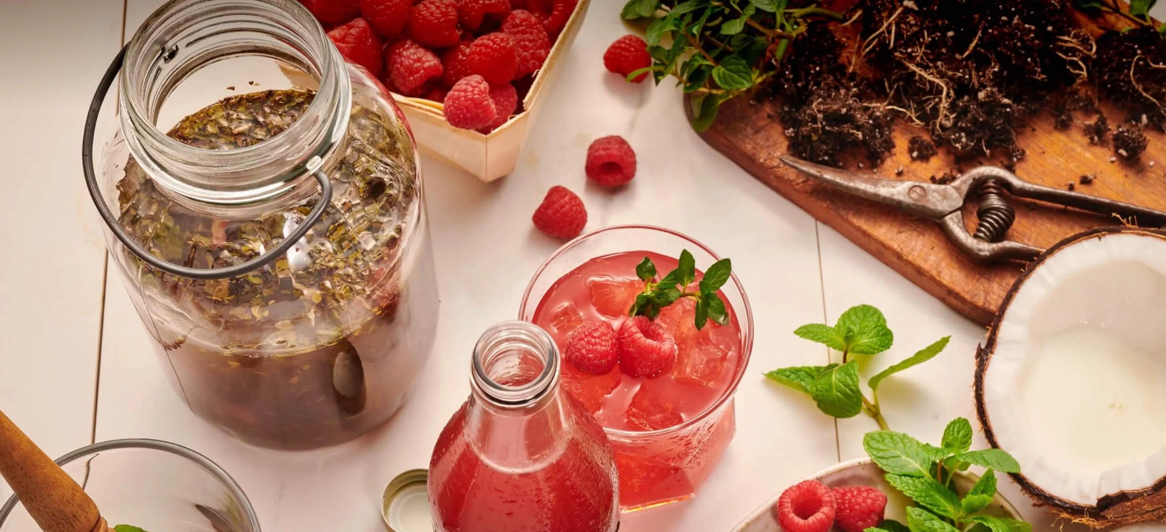 Glass jar with herbs, bottle of red drink, and raspberries on a white surface