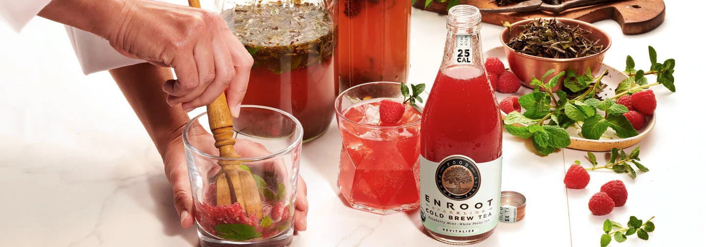 Person preparing a drink with raspberries and mint, surrounded by a bottle of Enroot Cold Brew Tea and other ingredients on a white surface.