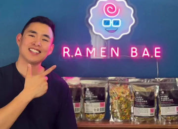 ramen bae founder pointing to sign in front of 'Ramen Bae' products with a neon sign in the background.
