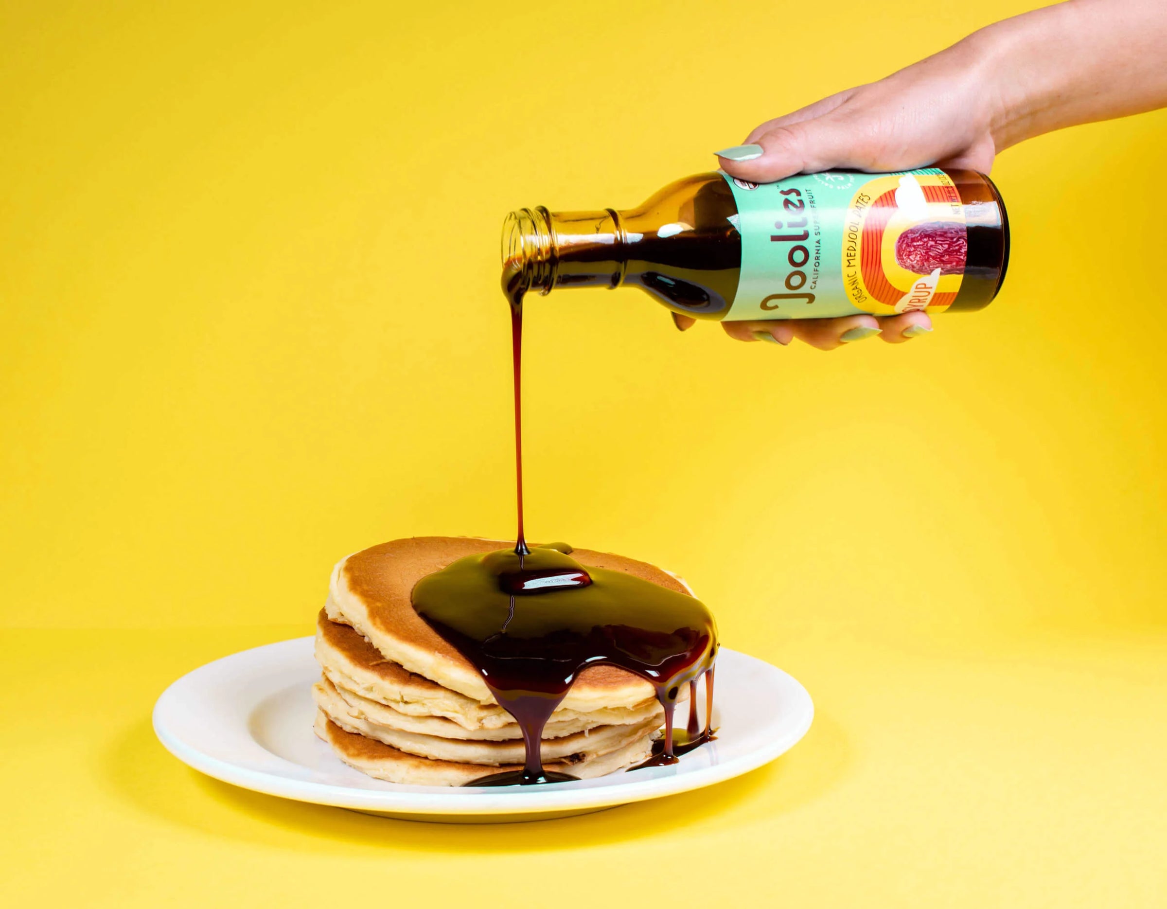 Person pouring syrup from a bottle labeled 'Joolies' onto pancakes on a yellow background