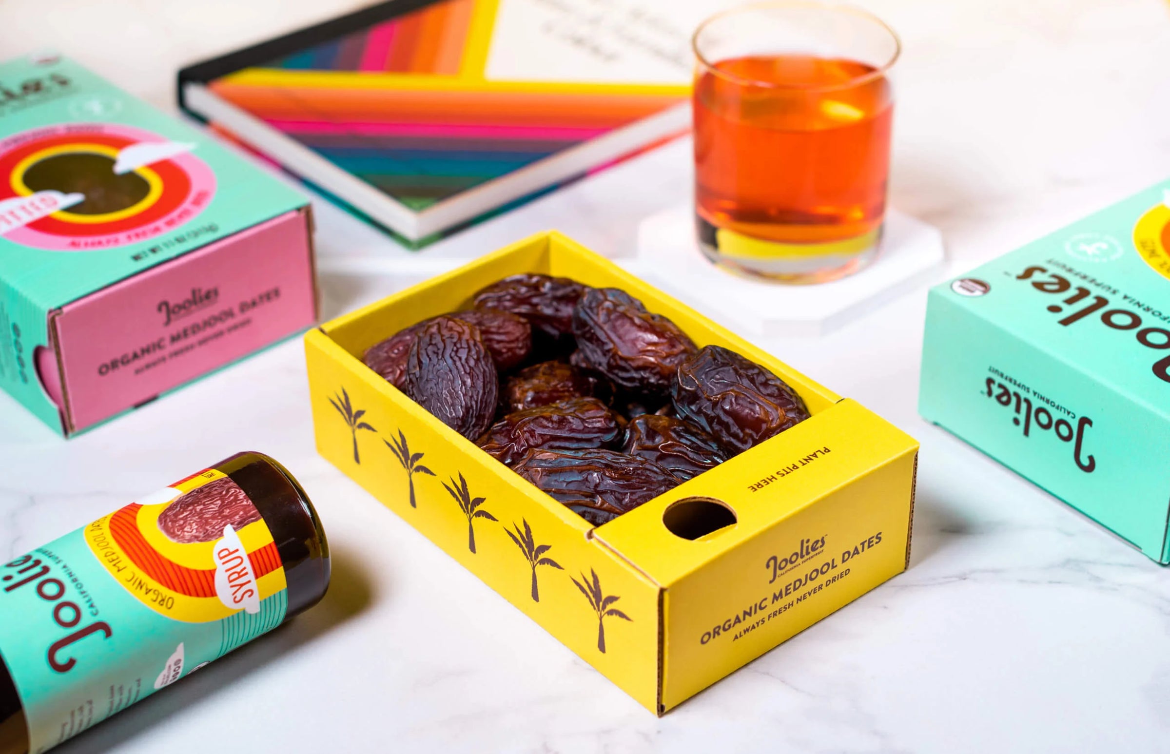 Box of Joolies organic medjool dates with colorful packaging and a glass of tea in the background.