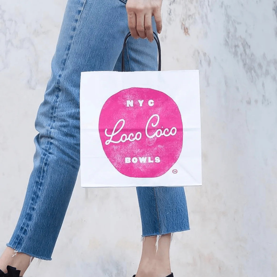 Person holding a white tote bag with pink logo and text, against a light gray background