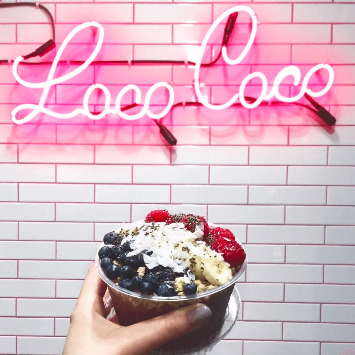 Hand holding a bowl of fruit salad in front of a pink neon sign with 'Loco Coco' on a tiled wall.