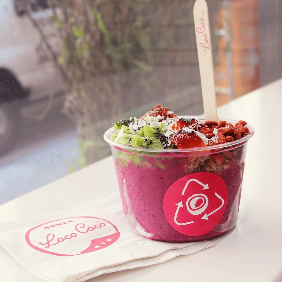 Pink smoothie bowl with toppings in a clear container on a white napkin, with 'Loco Coco' branding.