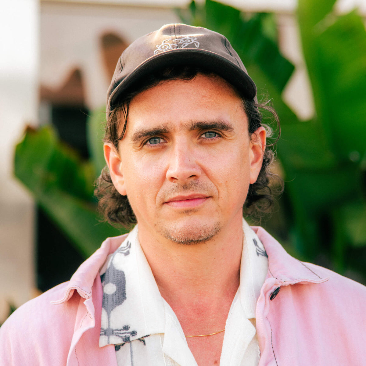 A close-up portrait of a man wearing a dark brown baseball cap, a light pink button-up shirt, and a white patterned collared shirt, set against a blurred background of green tropical leaves.