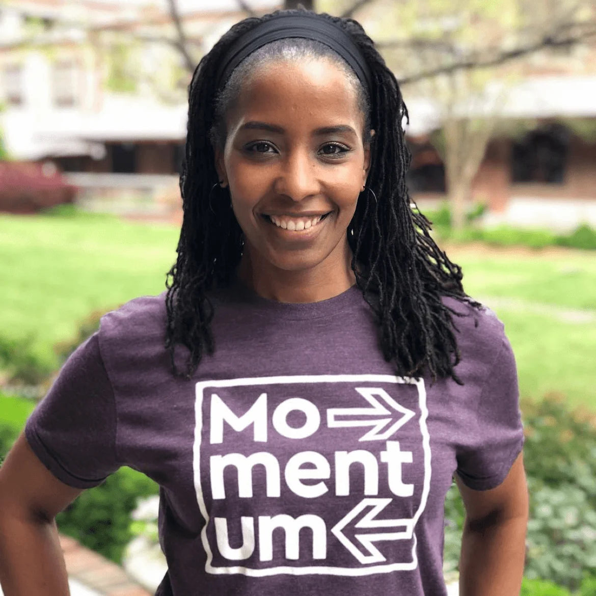 Person wearing a purple t-shirt with 'Momentum' text and arrows outdoors.