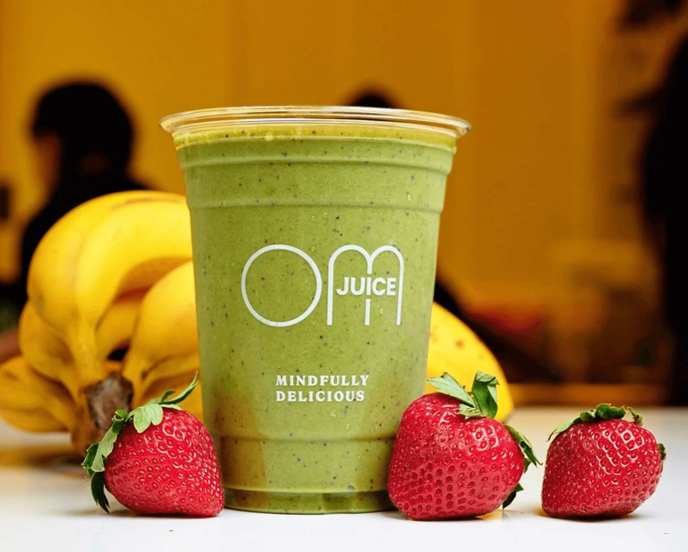 Green smoothie in a cup labeled 'OM Juice' with strawberries and bananas on a blurred background