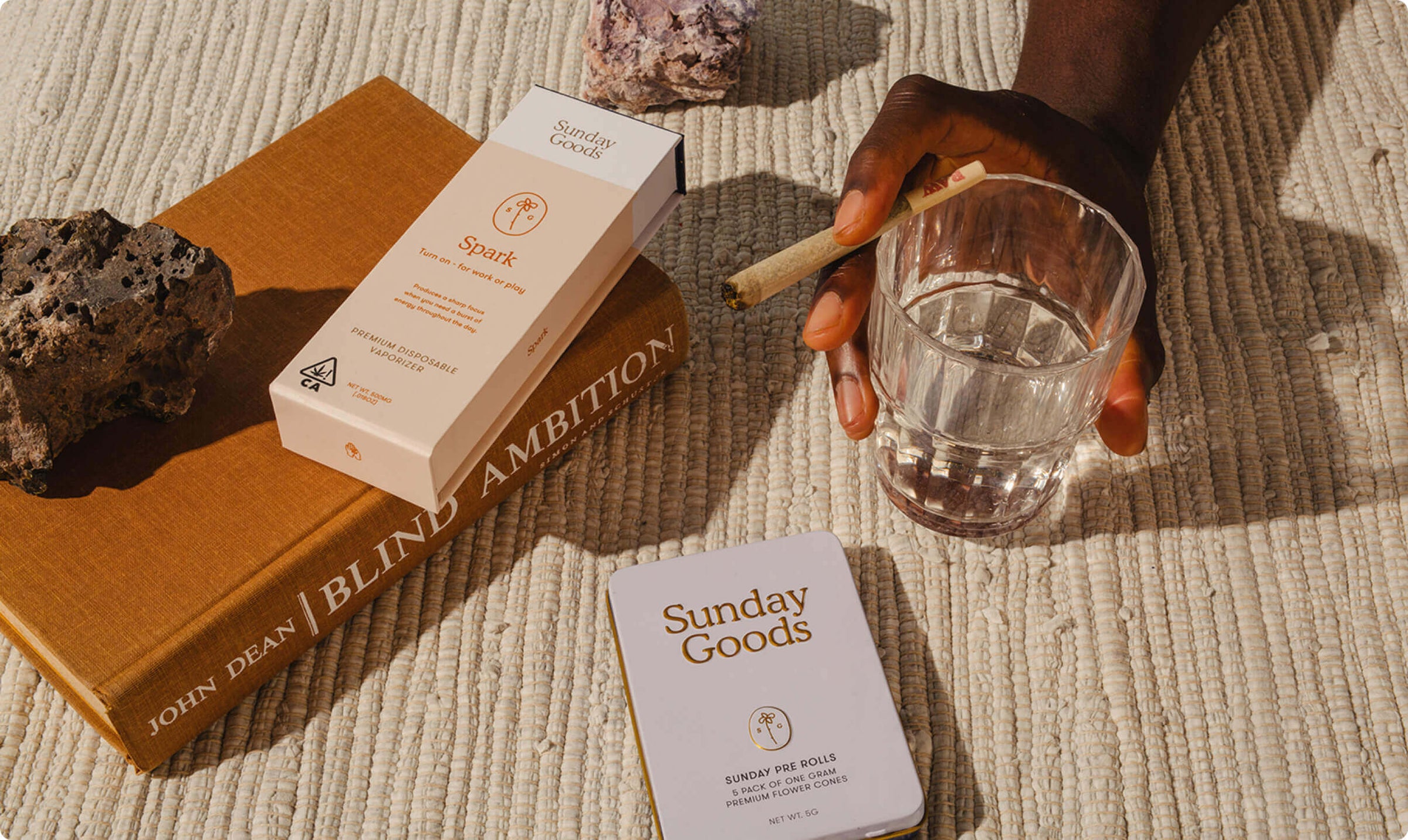 Hand holding a glass of water with 'Sunday Goods' product and books on a textured surface