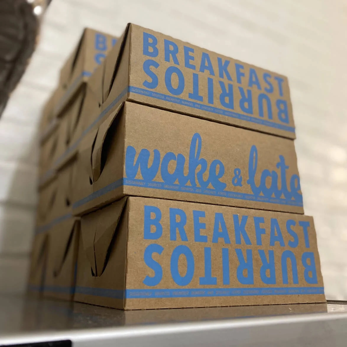 Stack of cardboard boxes with 'Breakfast Burritos' and 'Wake & Late' text on a neutral background