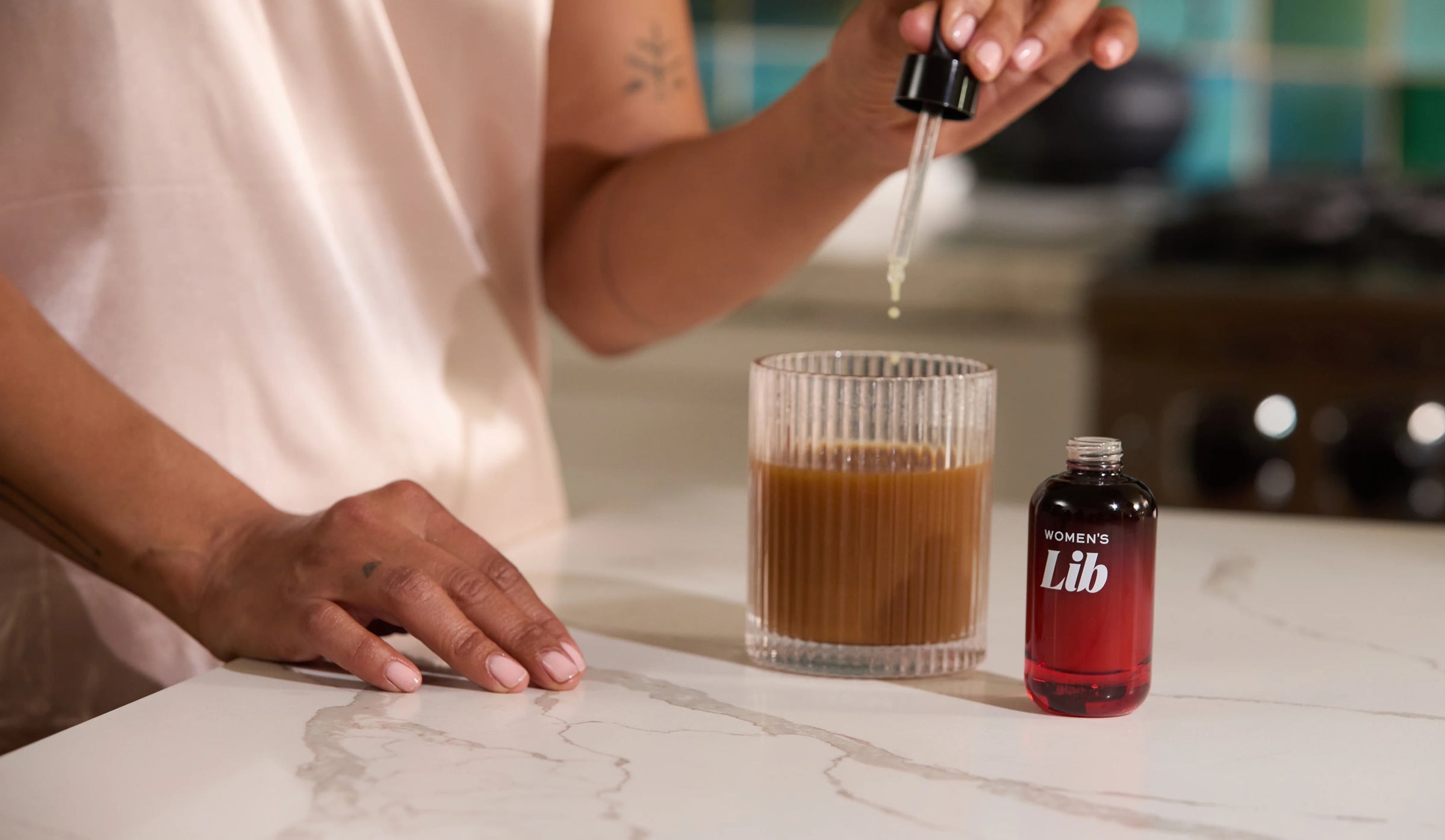Person adding a liquid from a dropper into a glass of brown liquid on a marble countertop.
